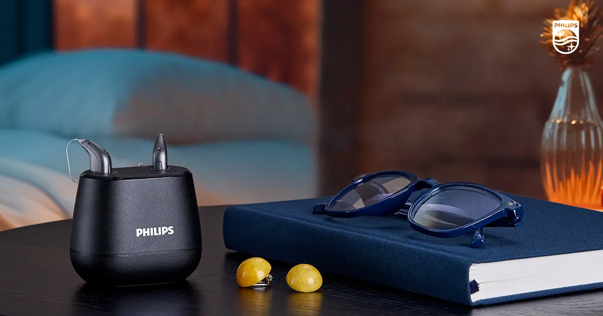 מטען Philips ובו זוג מכשירי שמיעה, על שידה בבית לצד ספר ומשקפיים